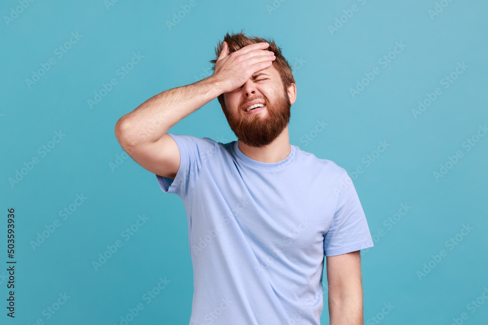 Portrait of upset sad bearded man standing with facepalm gesture, blaming himself, feeling sorrow regret because of bad memory. Indoor studio shot isolated on blue background. Portrait of upset sad bearded man standing with facepalm gesture, blaming himself, feeling sorrow regret because of bad memory. Indoor studio shot isolated on blue background.