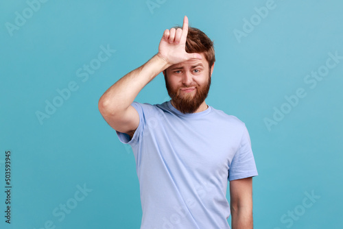 Portrait of handsome you g adult bearded man showing looser gesture holding fingers near forehead, sad because of silly mistake. Indoor studio shot isolated on blue background.