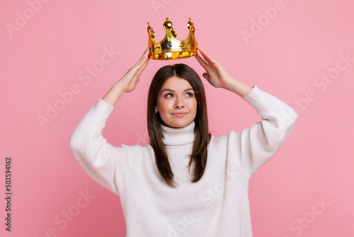 Bild auf Leinwand Young brunette woman putting on golden crown, looking with arrogance and smile, privileged status, wearing white casual style sweater