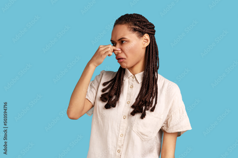 Stink, awful odor. Woman with black dreadlocks pinching nose, stop breathing bad odor, disgusted by smell of farting, her grimace expressing repulsion. Indoor studio shot isolated on blue background. Stink, awful odor. Woman with black dreadlocks pinching nose, stop breathing bad odor, disgusted by smell of farting, her grimace expressing repulsion. Indoor studio shot isolated on blue background.