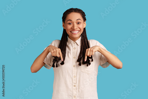 Gorgeous smiling woman with black dreadlocks pointing down, advertises new product, looking at camera with happy expression, wearing white shirt. Indoor studio shot isolated on blue background.