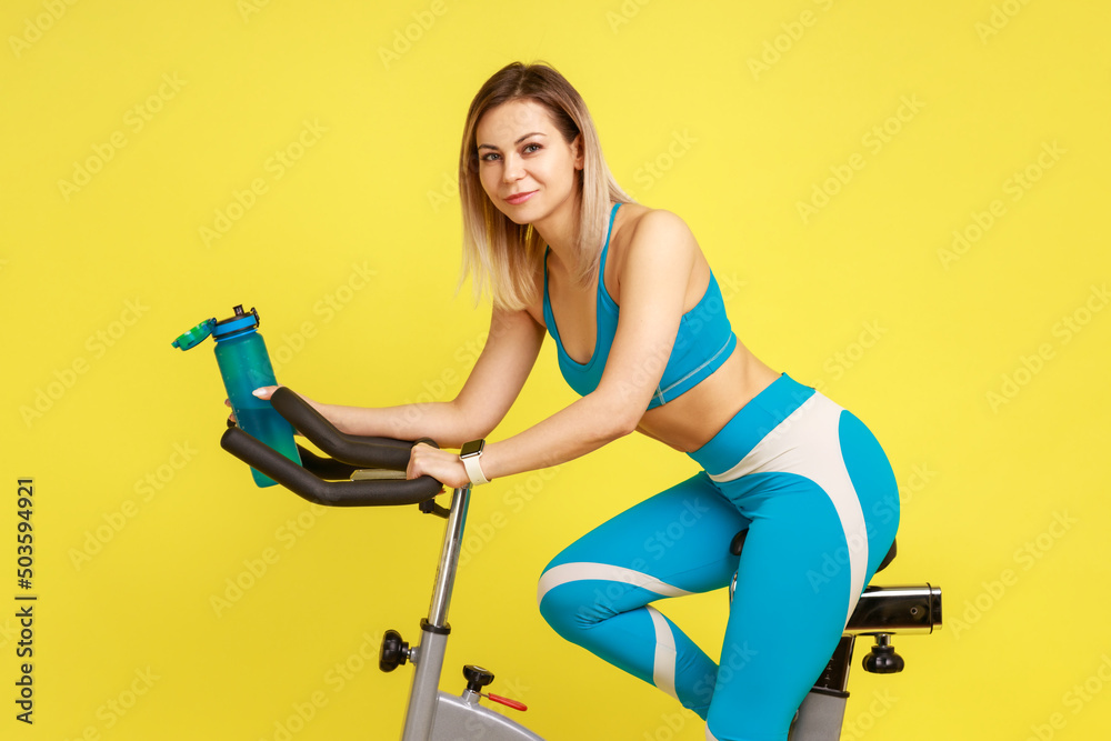 Side view of portrait of blonde athletic woman on exercise bike with bottle of water in hands, fitness and diet concept, wearing blue sportswear. Indoor studio shot isolated on yellow background. Side view of portrait of blonde athletic woman on exercise bike with bottle of water in hands, fitness and diet concept, wearing blue sportswear. Indoor studio shot isolated on yellow background.