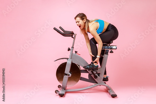 Full length portrait of young adult sportswoman was injured riding a home bike, holding her leg and writhes in pain, wearing sports tights and top. Indoor studio shot isolated on pink background. photo