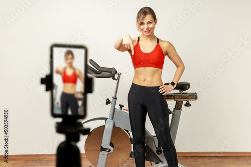 Smiling sporty woman blogger in top standing near exercise bike, recording video for sport vlog, pointing finger down, asking to subscribe. Indoor studio shot on gray wall background.