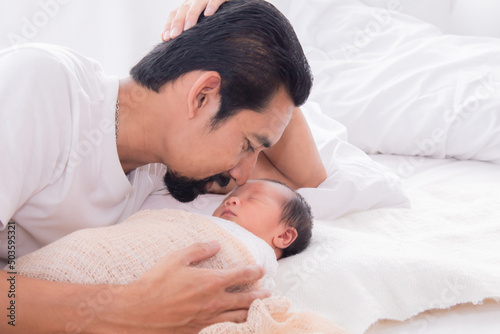 Close up of Asian young father kissing newborn while baby deeply sleeping with happiness. Little infant wrapped in thin white cloth with happy and safe. Dad and toddler spend time together.