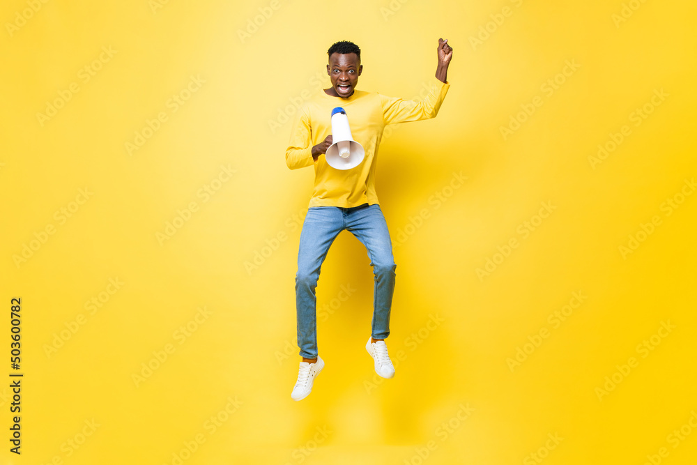 Jumping portrait of young African man yelling on megaphone in isolated yellow color background