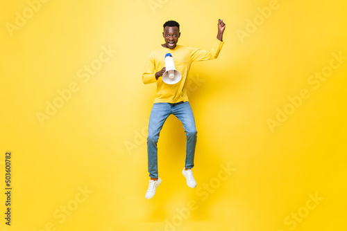 Jumping portrait of young African man yelling on megaphone in isolated yellow color background © Atstock Productions