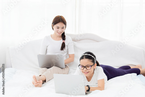Slective focus Beautiful young Asian mother working on laptop. Teenagers doing homework next to their babysitter. Two sisters spend time together in bed. Mom teaching daughter homework.
