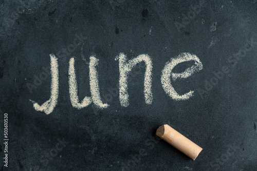 June. Name of the first summer month written in yellow chalk on a black chalkboard. Handwritten text. A piece of colored chalk lies next to it. photo