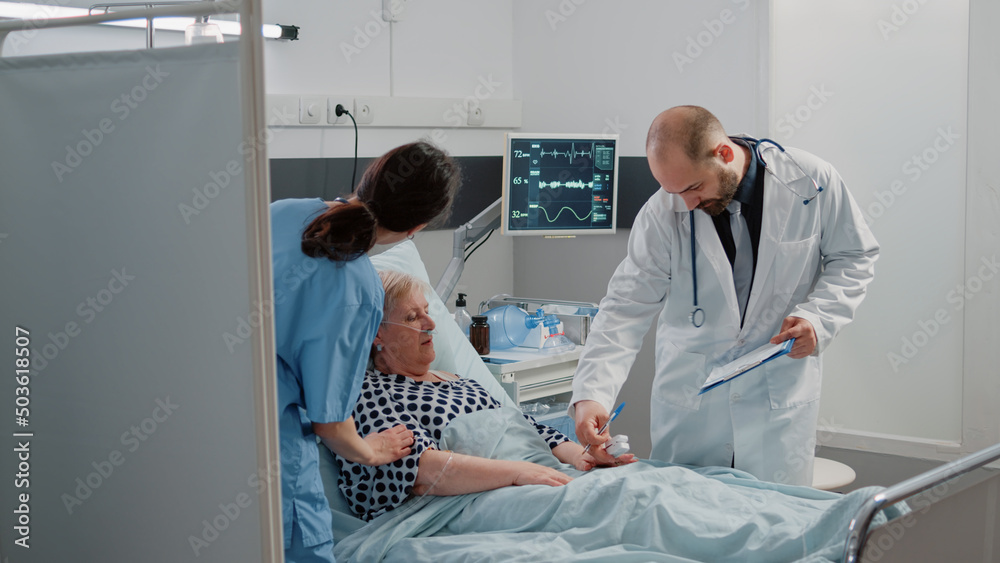 Fototapeta premium Ill patient discussing disease and treatment with doctor in hospital ward. Retired woman with nasal oxygen tube and IV drip bag talking to medic at checkup visit while nurse giving support
