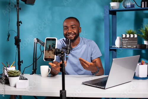 Smiling vlogger talking with audience in front of recording smartphone during online live show sitting at desk. Content creator interacting with fanbase in studio looking at live video podcast setup. photo