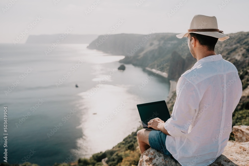 Digital nomad, man in the hat, a businessman with a laptop sits on the rocks by the sea during sunset, makes a business transaction online from a distance. Remote work on vacation.