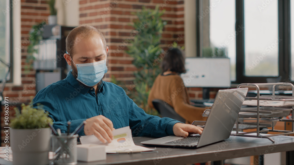 Employee wearing face mask and analyzing charts papers while working with laptop on business project. Entrepreneur using technology and files to work on presentation in startup office. Employee wearing face mask and analyzing charts papers while working with laptop on business project. Entrepreneur using technology and files to work on presentation in startup office.