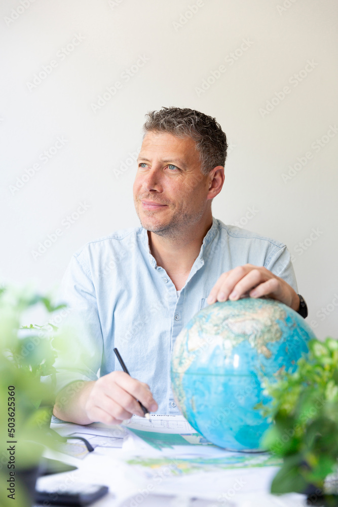 Business portrait of environmental engineer, ecologist, garden architect sitting at his work table holding a globe in his hands and surrounded by plants Business portrait of environmental engineer, ecologist, garden architect sitting at his work table holding a globe in his hands and surrounded by plants