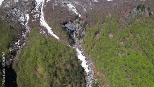 Rragam Waterfall on slope of rocky mountain surrounded by snow and green wild forest in Albania photo