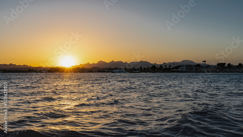 Sunset on the Red Sea. The rays of the setting sun illuminate a picturesque mountain range. The sky is colored orange. Waves on the water. Egypt. Safaga
