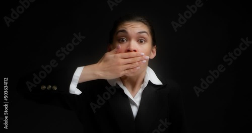 Funny brunette, dressed in formal business clothes, stands on an isolated black background in the studio, covers her mouth with her hands, blurted out something wrong, looks around with her eyes photo