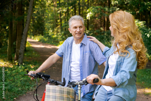 Happy mature couple on bike ride on sunny day on path in forest. Joyful pastime of Caucasian pensioners in casual comfortable clothes for a walk