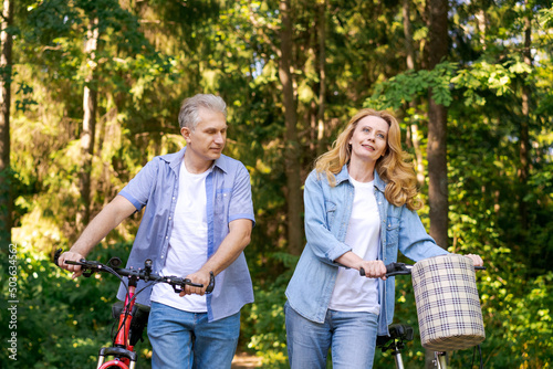 Active old age, people and lifestyle concept happy senior couple fixing bike, talking on summer city park on path in sunny forest, wearing casual clothes cute caucasian man and woman