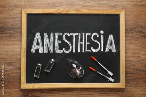 Blackboard with word Anesthesia, vials, mask and syringes on wooden table, top view