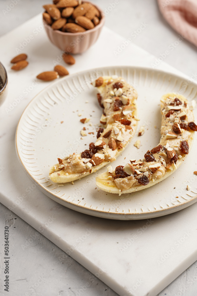 Two banana halves ready with peanut butter topping, raisins, almonds and chia seeds on white plate on marble board Two banana halves ready with peanut butter topping, raisins, almonds and chia seeds on white plate on marble board