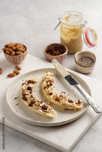 Two banana halves ready with peanut butter topping, raisins, almonds and chia seeds on white plate on marble board