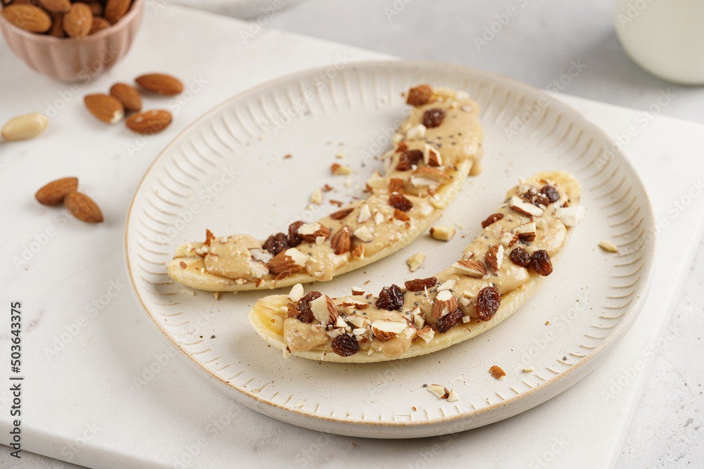 Two banana halves ready with peanut butter topping, raisins, almonds and chia seeds on white plate on marble board Two banana halves ready with peanut butter topping, raisins, almonds and chia seeds on white plate on marble board