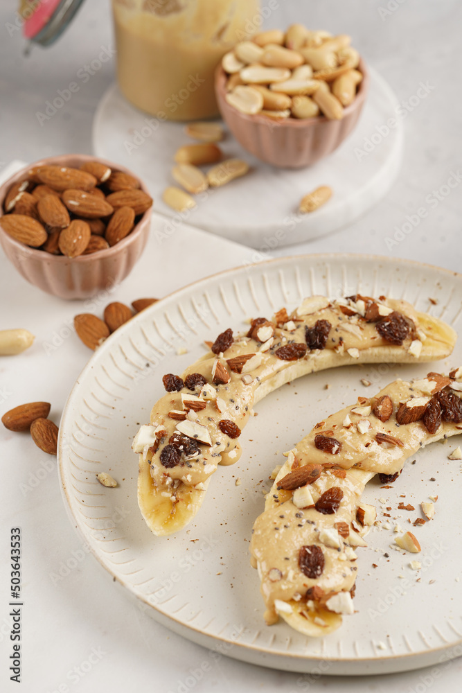 Two banana halves ready with peanut butter topping, raisins, almonds and chia seeds on white plate on marble board Two banana halves ready with peanut butter topping, raisins, almonds and chia seeds on white plate on marble board