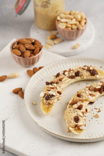 Two banana halves ready with peanut butter topping  raisins  almonds and chia seeds on white plate on marble board