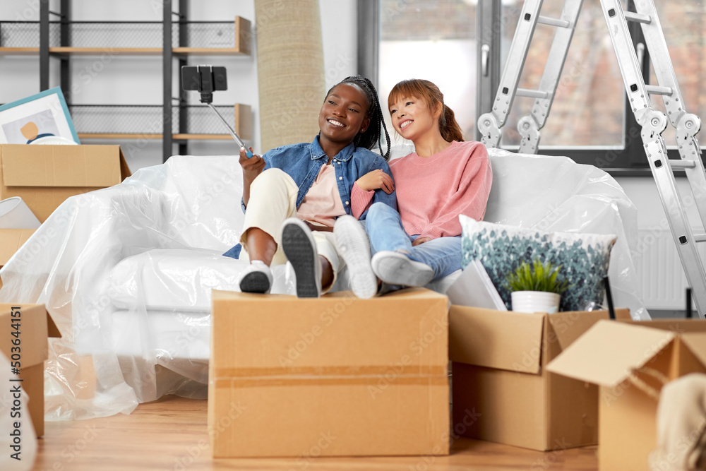 moving, people and real estate concept - happy smiling women with smartphone and selfie stick taking picture at new home moving, people and real estate concept - happy smiling women with smartphone and selfie stick taking picture at new home