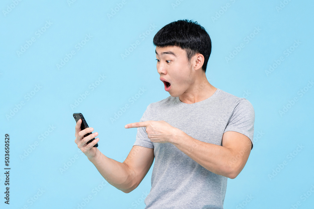 Amazed Asian man in casual wear pointing at cellphone while standing on light blue isolated background in studio Amazed Asian man in casual wear pointing at cellphone while standing on light blue isolated background in studio