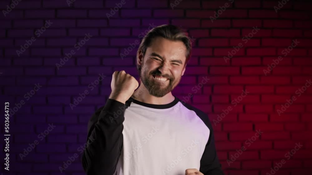 Portrait of satisfied young guy making yes gesture with arms raised while posing in studio with neon light around. Concept of people, lifestyles and sincere emotions. Portrait of satisfied young guy making yes gesture with arms raised while posing in studio with neon light around. Concept of people, lifestyles and sincere emotions.