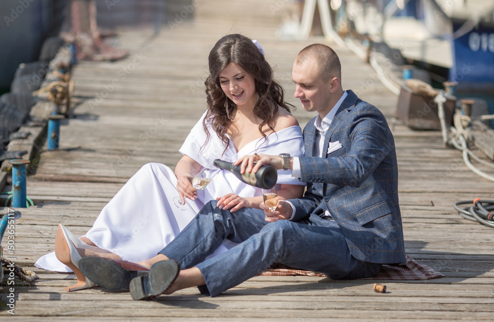 a young man in a suit and a woman in a white dress are sitting on a pier by the sea and drinking champagne, they are having a wedding. a young man in a suit and a woman in a white dress are sitting on a pier by the sea and drinking champagne, they are having a wedding.