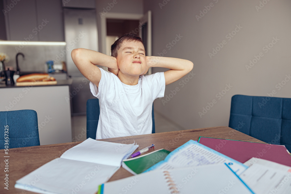 Caucasian small lazy schoolboy closing ears with hands at homework and textbooks. Caucasian small lazy schoolboy closing ears with hands at homework and textbooks.