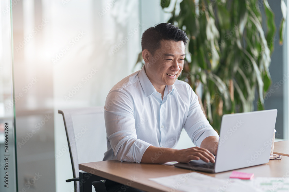 Smiling Asian businessman working on laptop at her workplace in modern office. Business concept Smiling Asian businessman working on laptop at her workplace in modern office. Business concept