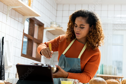 African American concentrated woman vlogger with curly hair mixing glaze in bowl with whisk while showing recipe in kitchen online photo