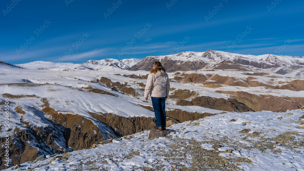Naklejka premium A man in a down jacket stands on a high-altitude plateau at the edge of the gorge and looks into the distance. Back view. The red-brown slopes of the mountains are snow covered. Blue sky. Altai Mars