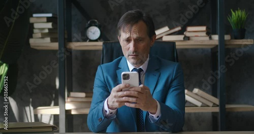 Shocked Senior Business Man Reacting on Bad Message on Smartphone reading terrible new while sitting at office workplace. Male is shocked by what he saw on his mobile phone