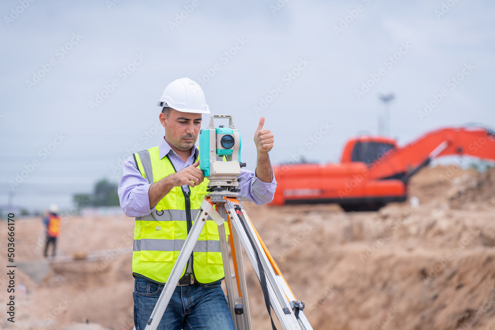 Surveyor engineer wearing safety uniform ,helmet and radio communication with equipment theodolite to measurement positioning on the construction site of the road with construct machinery background Surveyor engineer wearing safety uniform ,helmet and radio communication with equipment theodolite to measurement positioning on the construction site of the road with construct machinery background