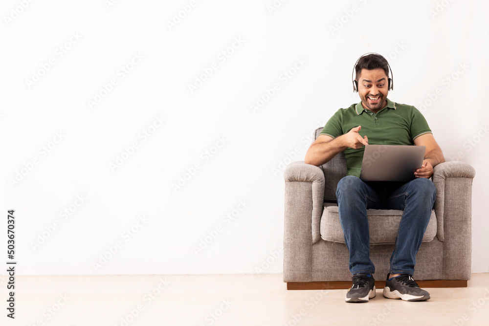 Cheerful young man listening music on headphones and using laptop while sitting on sofa