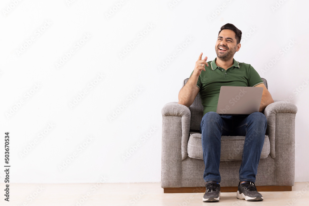 Portrait of a cheerful young man pointing elsewhere while sitting on sofa with laptop on his lap