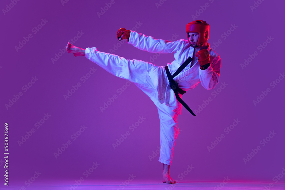 Studio shot of male taekwondo fighter in sports uniform in action isolated over purple background in neon light. Concept of sport, workout, competition, ad