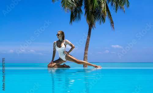 Elegant tanned woman in white swimsuit in pool on tropical Maldives island. Beautiful bikini body girl in pool with view on horizon. Sexy model near the pool on beautiful Indian ocean landscape. 