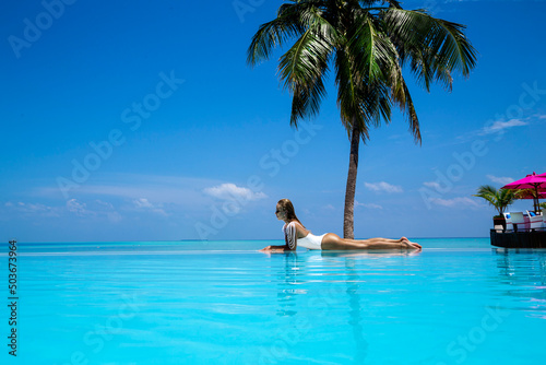 Elegant tanned woman in white swimsuit in pool on tropical Maldives island. Beautiful bikini body girl in pool with view on horizon. Sexy model near the pool on beautiful Indian ocean landscape. 