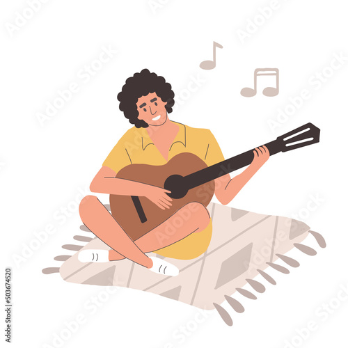 A young man is sitting on a blanket and playing the guitar. The guy smiles sings songs and plucks the strings with his legs folded on a white background