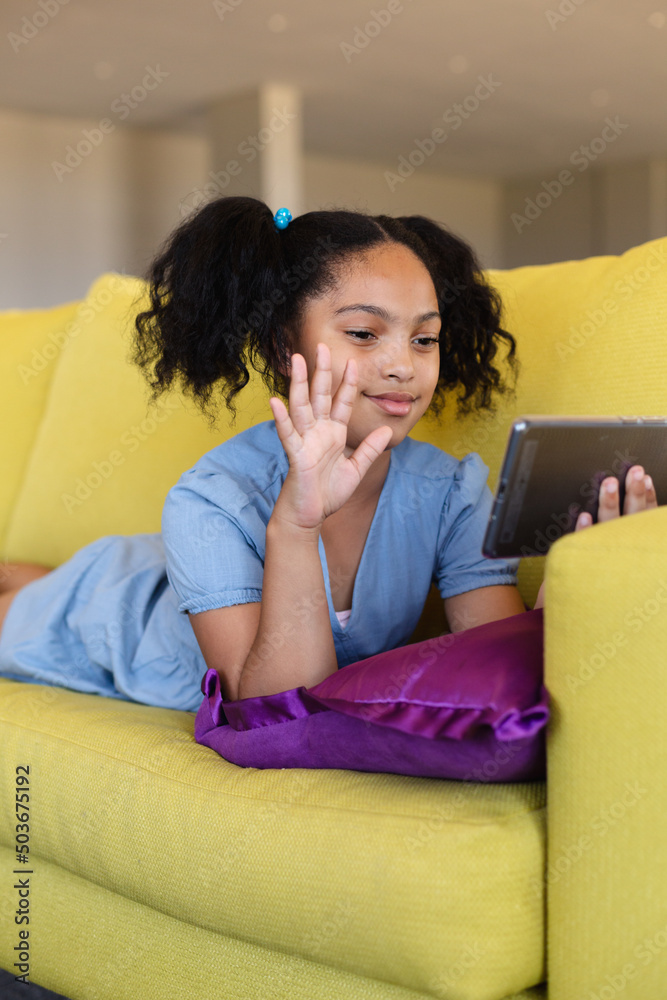 Biracial elementary girl waving over smart phone during video call while lying on couch in school Biracial elementary girl waving over smart phone during video call while lying on couch in school