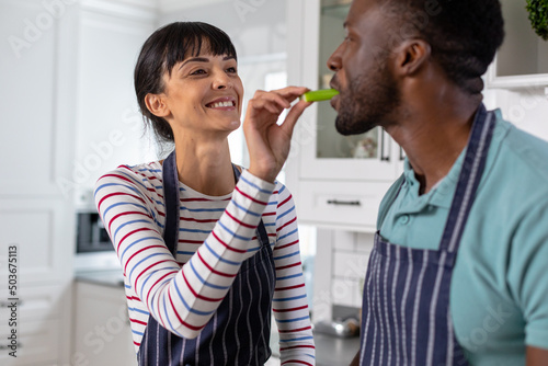 Smiling caucasian young woman feeding vegetable to african american boyfriend at home