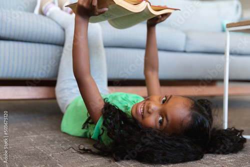 Portrait of african american elementary girl reading book while lying on floor with legs over couch