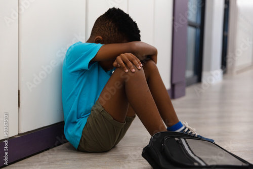 Sad african american elementary schoolboy hugging knees while sitting on floor in corridor © wavebreak3