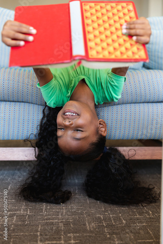 Smiling african american elementary girl reading book while lying upside down on couch in school